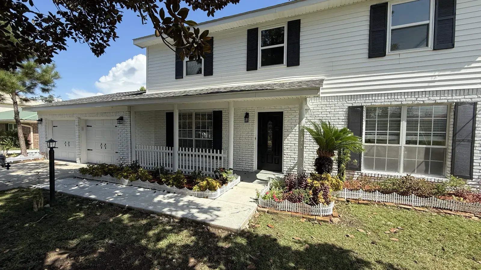 Finished two-story home with white painted brick, white siding, black shutters, a covered front porch and fresh landscaping