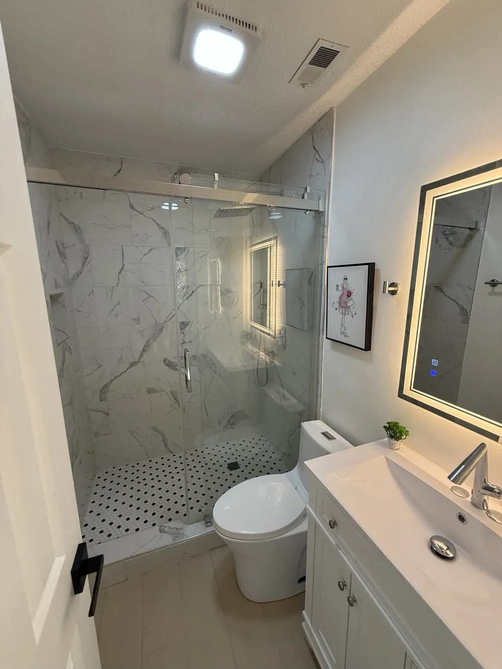 Guest bathroom with a marble-look tiled shower, frameless sliding glass door, black and white penny tile pan and a backlit mirror