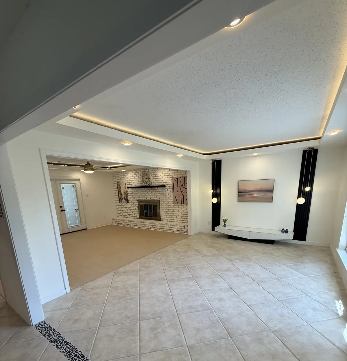 Open living room with a whitewashed brick fireplace, recessed cove lighting, tile floors and a floating media console