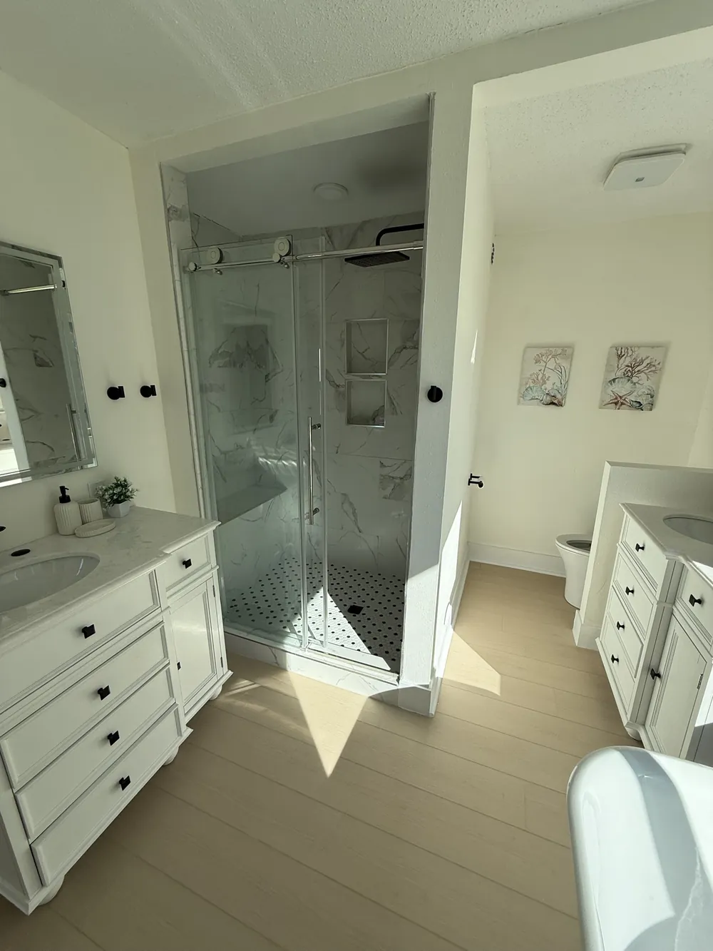 Primary bathroom with twin white vanities, black hardware, a marble-look tiled shower with frameless glass and a soaking tub