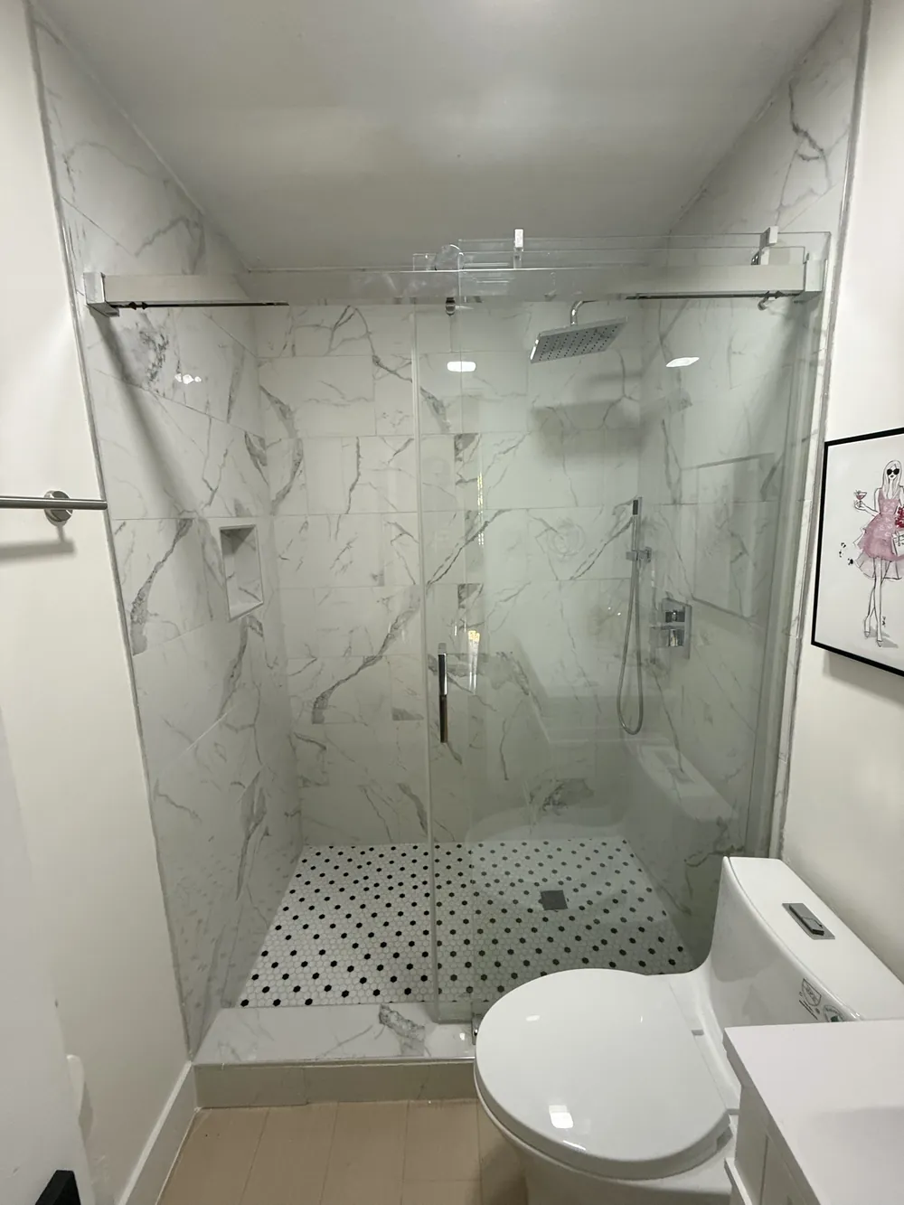 Walk-in shower tiled in large marble-look porcelain with a frameless sliding glass door, rain head and black and white penny tile pan